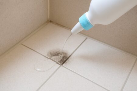 風呂の床の隅に液体の漂白剤が透明な状態で撒かれている場面。床はベージュ系のタイルで、窓からの自然光が差し込み、淡い陰影が見える清潔感のある実写構図。